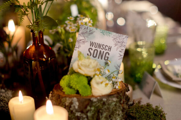 natürliche Hochzeitstischdeko mit Blumen, Blättern, Moos und Sukkulenten, in die eine Karte mit der Aufschrift Wunschsong gesteckt wurde