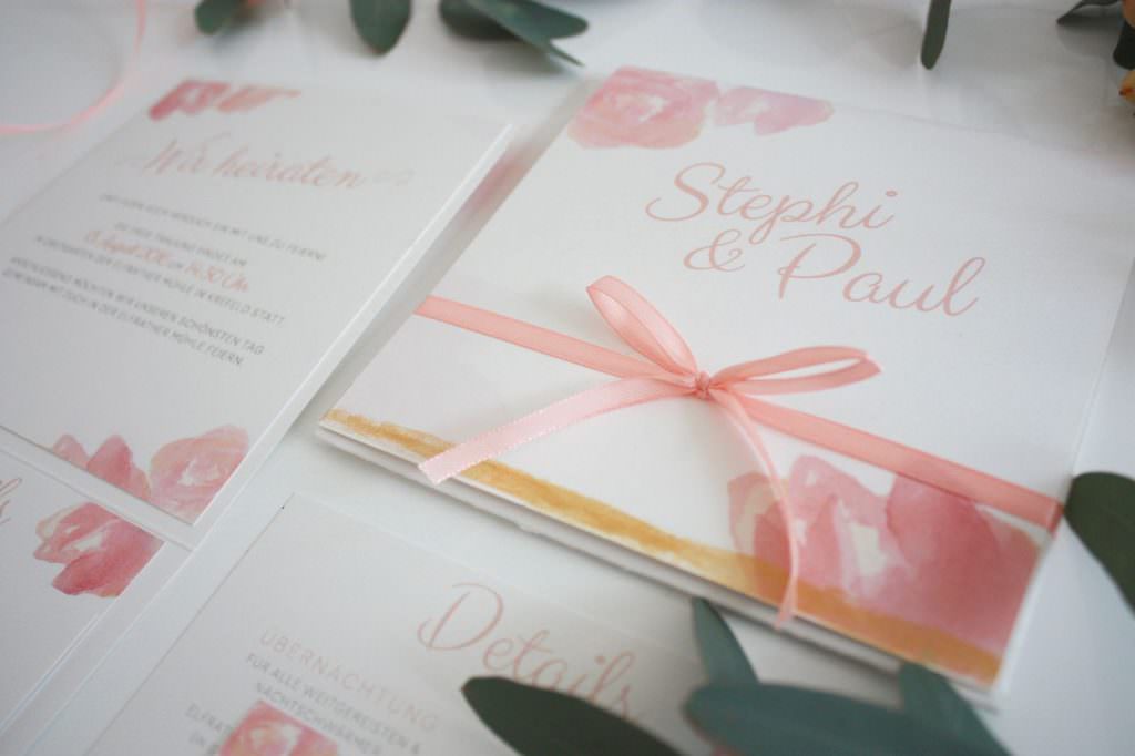 Weiße Hochzeitspapeterie mit Schrift, Verzierung und Schleife in Rosa