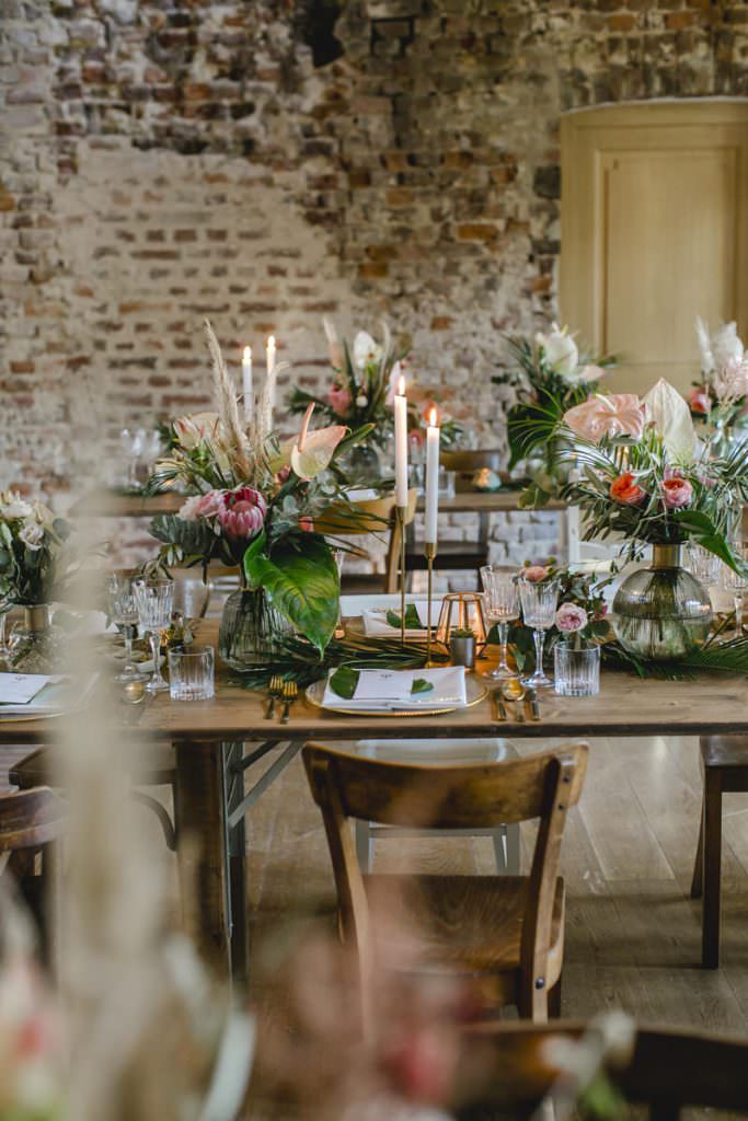 Hochzeitstischdeko mit viel Greenery, Palmenblättern und Protea, Flamingoblumen und Rosen in Rosa.