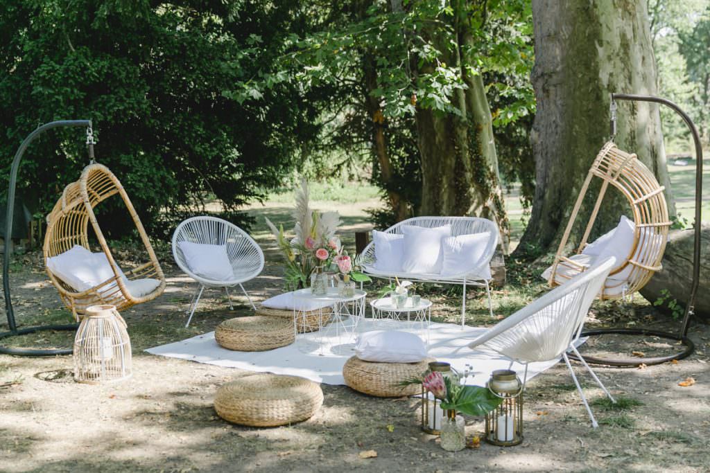 Lounge Ecke bei einer Outdoor Hochzeit mit weißen Acapulco Chairs, Rattan-Hängesesseln und Rattan Bodenhockern