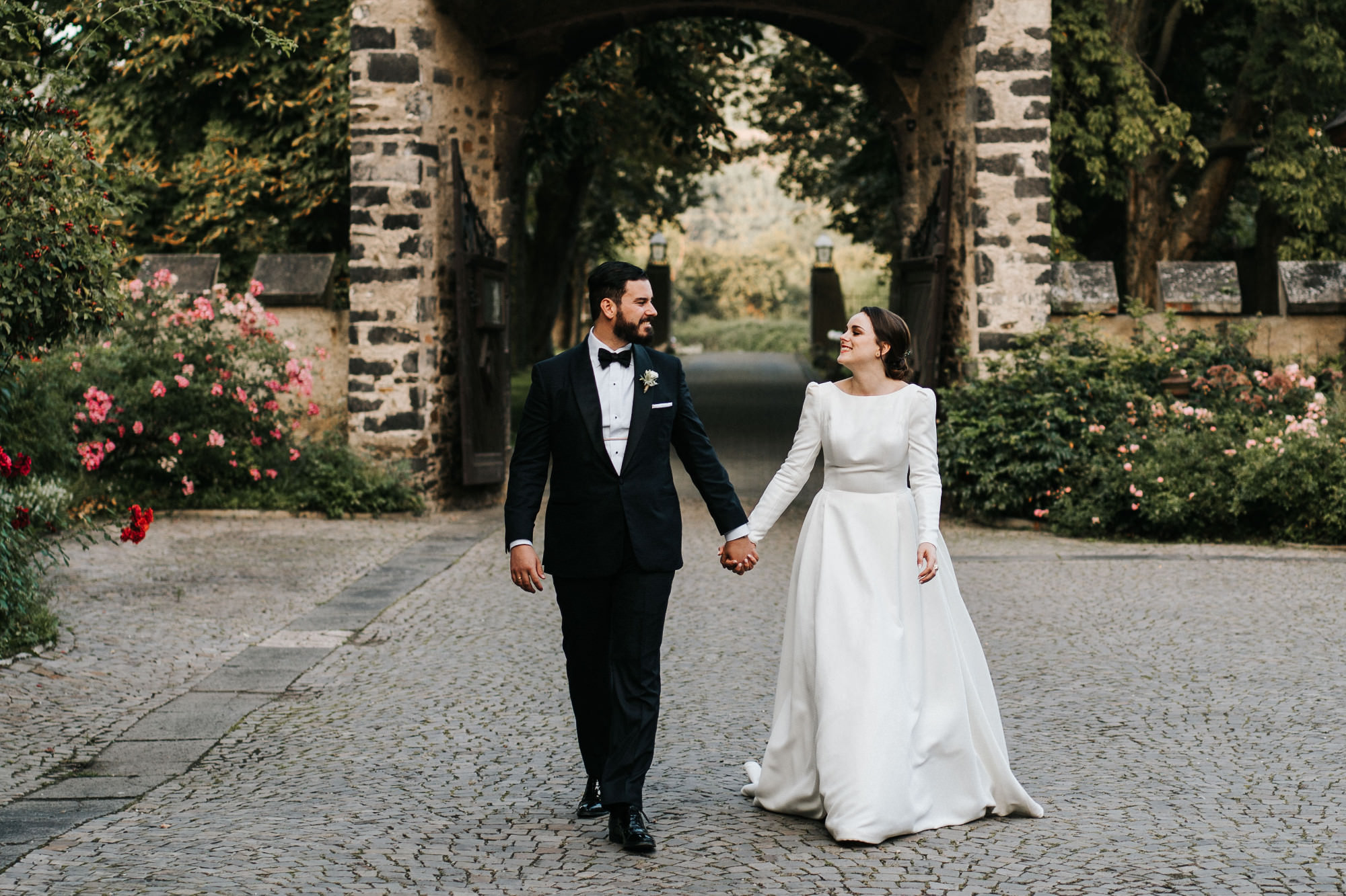 Ein Brautpaar geht Hand in Hand nach vorne ins Bild. Sie schauen sich an. Im Hintergrund eine Burgmauer