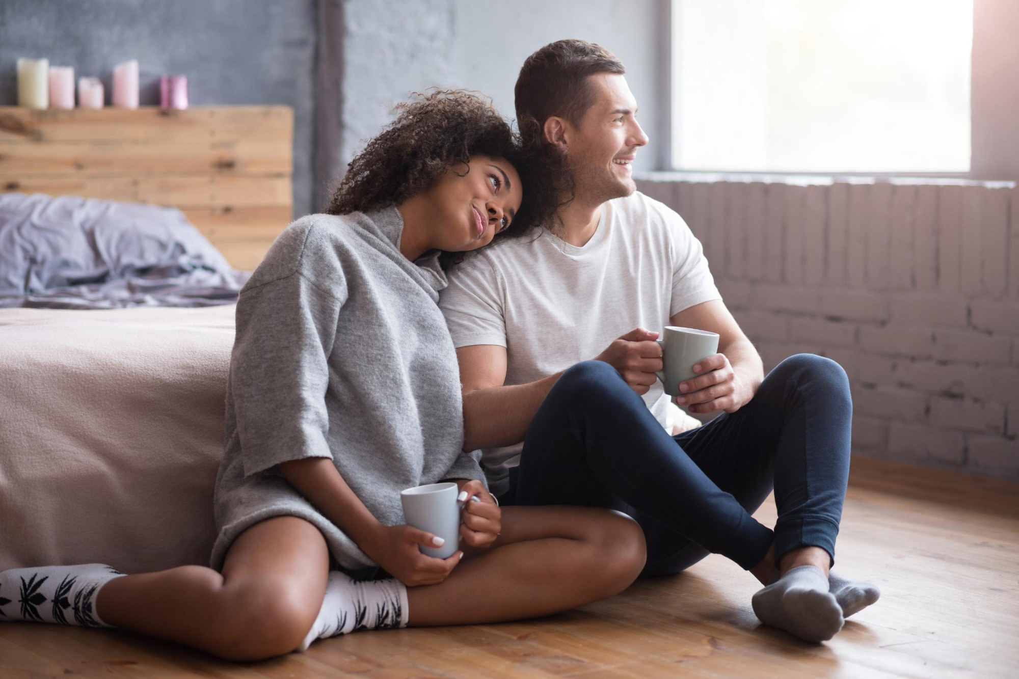 Ein Paar sitzt mit Kaffeetassen in der Hand auf dem Boden vor einem Bett und schaut zum Fenster, während sie ihren Kopf auf seine Schulter gelegt hat