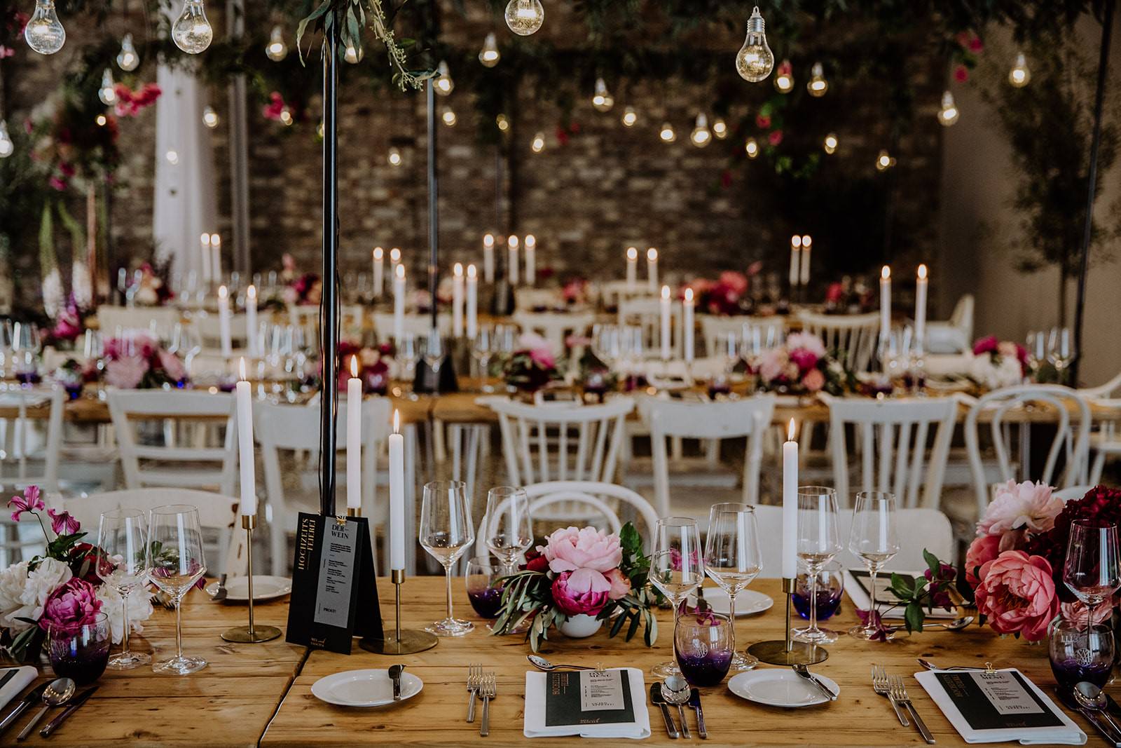 Ein Blick über lange Reihen von eingedeckten Hochzeitstischen. Der Raum wird festlich beleuchtet von Kerzen und Lichterketten.