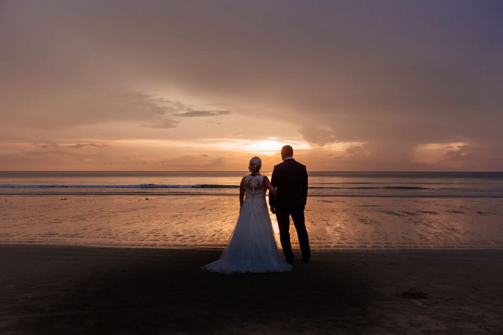 Ein Brautpaar steht am Strand und schaut in den Sonnenuntergang.