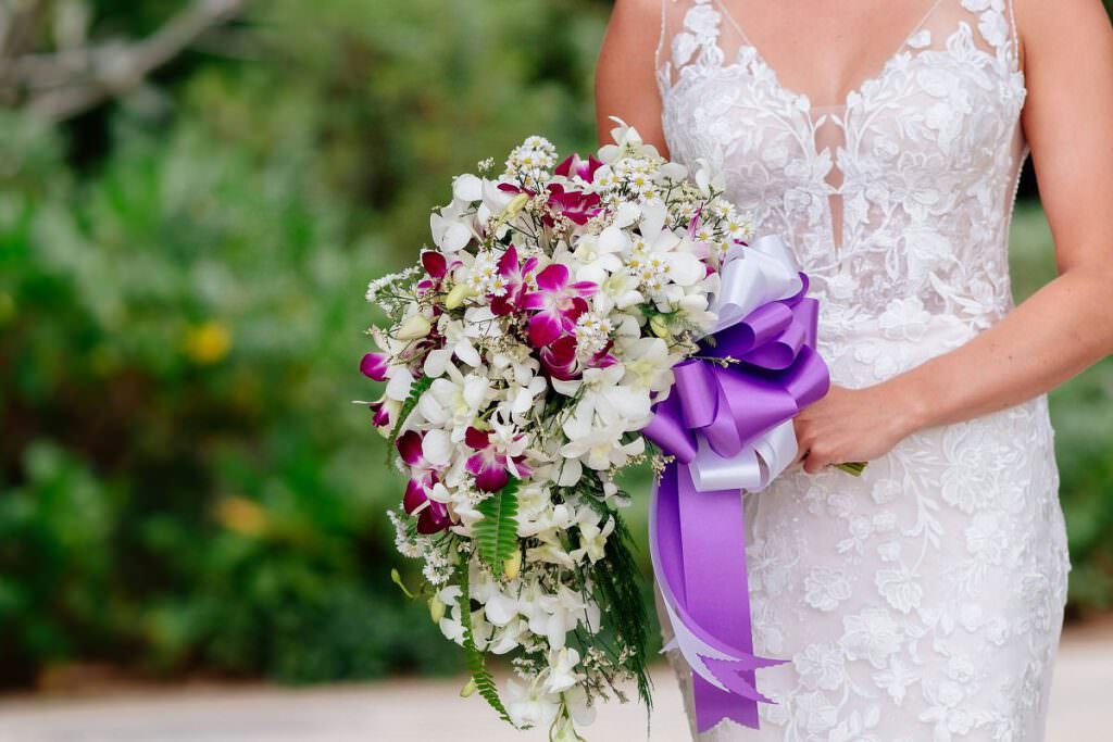 Eine Braut hält einen großen Brautstrauss in der Hand. Er ist mit einer lila Schleife zusammengebunden.