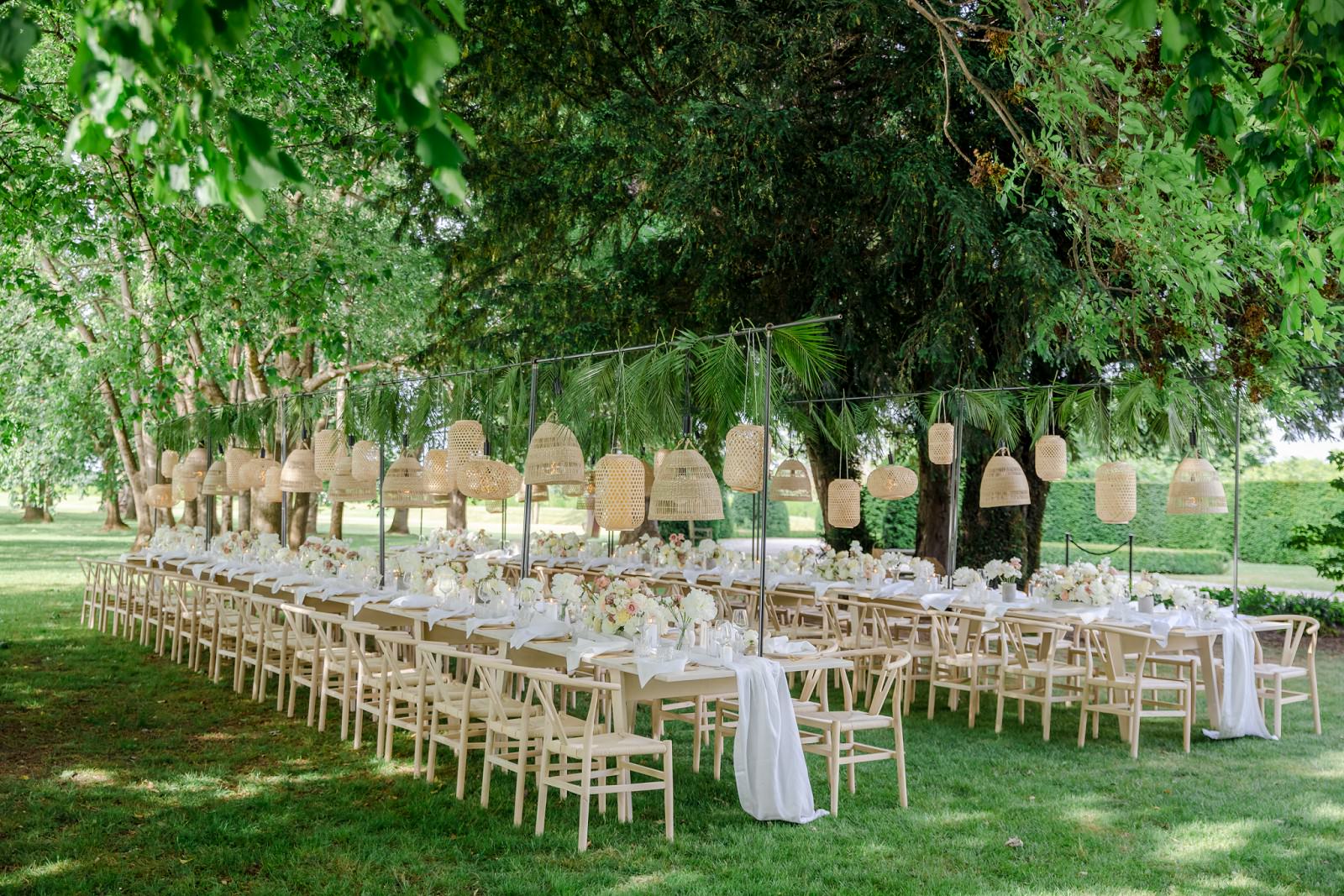 lange Hochzeitstafeln sind mit Lampenschirmen als Hängedeko unter Bäumen aufgestellt