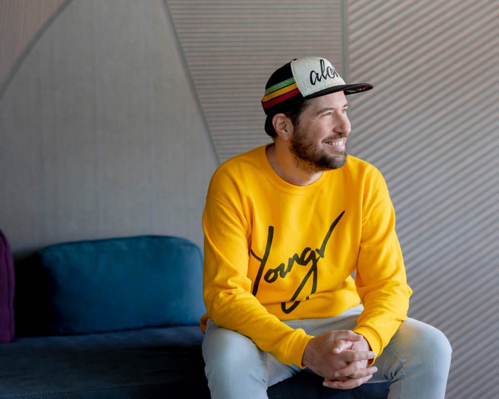 Hochzeits-DJ Markus Rosenbaum trägt einen gelben Youngr Pulli und sitzt auf einem blauen Sofa